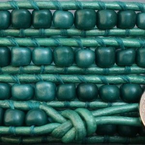 May include: Close-up of a teal wrap bracelet. The bracelet features square wooden beads woven into a teal leather band. A silver-colored button clasp secures the bracelet. The design is intricate and the color is vibrant.