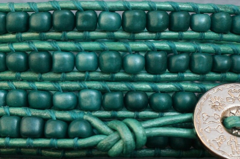 May include: Close-up of a teal wrap bracelet. The bracelet features square wooden beads woven into a teal leather band. A silver-colored button clasp secures the bracelet. The design is intricate and the color is vibrant.