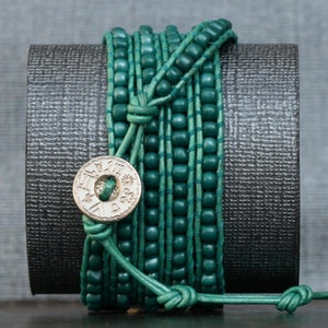 May include: A teal wrap bracelet with wooden beads and leather cord. The bracelet wraps multiple times around the wrist and features a decorative silver-colored button clasp with etched details. The bracelet is displayed on a dark gray surface.