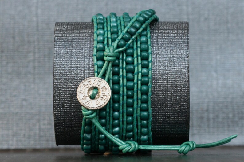 May include: A teal wrap bracelet with wooden beads and leather cord. The bracelet wraps multiple times around the wrist and features a decorative silver-colored button clasp with etched details. The bracelet is displayed on a dark gray surface.