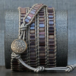 May include: A multi-layered bracelet with a gray leather base and purple glass beads. A decorative metal charm is attached to the leather strap.