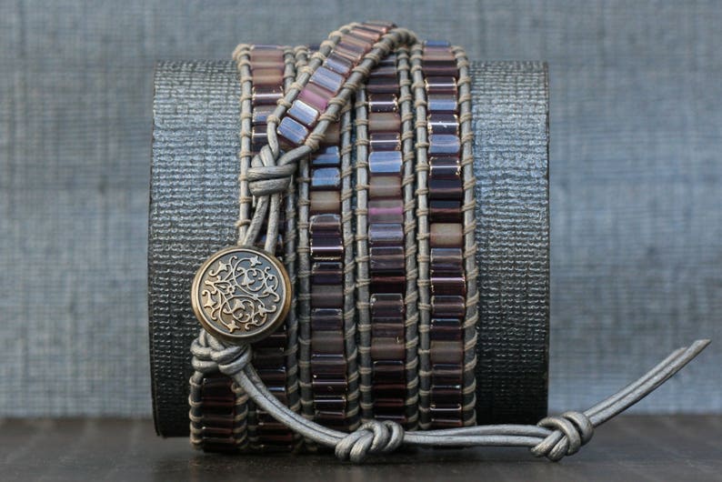 May include: A multi-layered bracelet with a gray leather base and purple glass beads. A decorative metal charm is attached to the leather strap.