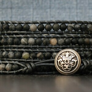 Peut inclure: Un bracelet multi-rangs en cordon de cuir noir et perles de pierre gris foncé et blanc. Un charme argenté avec un motif de couronne est attaché au bracelet.
