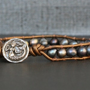 May include: A brown leather bracelet with a silver mermaid charm and dark gray pearls.