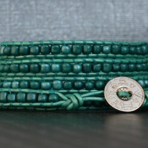May include: A wrap bracelet featuring teal-colored beads and leather. The bracelet wraps multiple times around the wrist and is secured with a silver-colored button clasp. The beads are square-shaped and evenly spaced.