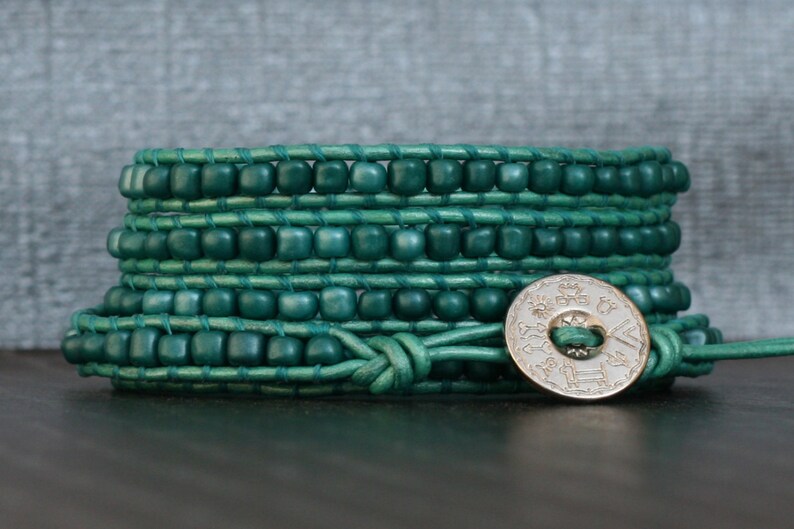 May include: A wrap bracelet featuring teal-colored beads and leather. The bracelet wraps multiple times around the wrist and is secured with a silver-colored button clasp. The beads are square-shaped and evenly spaced.