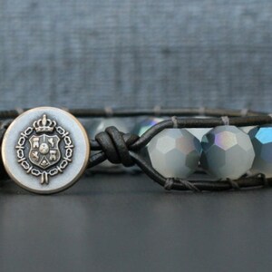 Könnte beinhalten: Ein schwarzes Lederarmband mit einem silbernen Knopf, der ein Kronen- und Schilddesign zeigt. Das Armband ist mit schimmernden grauen Perlen verziert.