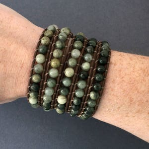 May include: A multi-strand bracelet with green and brown stones wrapped in brown leather. The bracelet is worn on a person's wrist.
