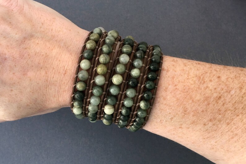 May include: A multi-strand bracelet with green and brown stones wrapped in brown leather. The bracelet is worn on a person's wrist.