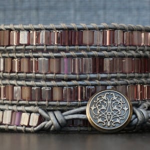 May include: A multi-layered bracelet made of brown leather and pink glass beads. The bracelet has a decorative metal clasp with a floral design.