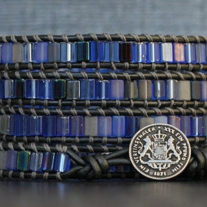 Puede incluir: Un brazalete de cuero de m&uacute;ltiples capas con cuentas de vidrio azul iridiscente y un dije de moneda de plata con el texto "EIN THALER - EIN PFUND 1871".