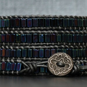 May include: A multi-layered bracelet with iridescent black beads and a silver charm with a swirling design. The bracelet is made with black leather and has a simple closure.