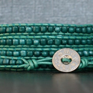 May include: A wrap bracelet featuring teal-colored wooden beads and leather cord. The bracelet wraps multiple times around the wrist and is secured with a decorative silver button clasp. The bracelet is a handmade accessory.