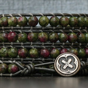 May include: A multi-layered leather wrap bracelet with green and pink gemstone beads and a silver charm with a cross design.