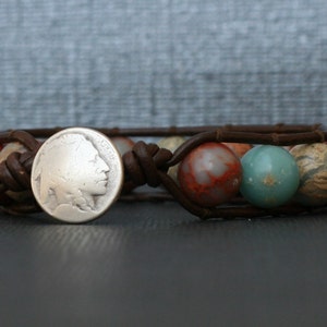 May include: A brown leather bracelet with a silver coin charm and colorful gemstone beads. The coin features a profile of a Native American man.