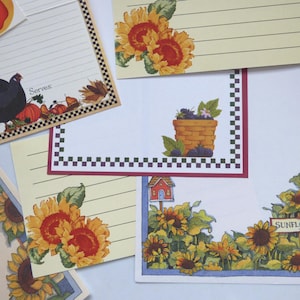 May include: Assortment of decorative recipe cards with various designs. Some cards feature sunflowers, a basket of berries, and a rooster with pumpkins. The cards have lined paper for writing recipes. A sticker of a red apple is also visible.