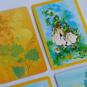 May include: Close-up of several tarot cards with yellow borders. One card features a yellow and orange background with plant silhouettes. Another card shows a whimsical illustration of figures in green clothing on a mushroom. The cards have a vintage aesthetic.