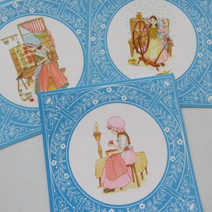 May include: Three blue cards with white floral borders and illustrations of girls spinning yarn. Each card features a different girl in a different pose.