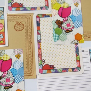 May include: A collection of stationery featuring a character in a pink bonnet holding a teddy bear, set against a colorful hexagonal patchwork background. Includes lined note cards with illustrations of a pitcher, apple, and flower.