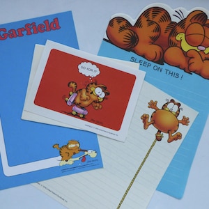 May include: A collection of Garfield-themed stationery items. Includes a blue notepad with "Garfield" in red, a red card with Garfield on a skateboard and the text "GO FOR IT", and a notepad with Garfield sleeping. The items feature the orange cat.
