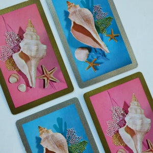 May include: Four playing cards with seashell and starfish designs. Each card features a gold border and a background of either pink or blue. Seashells, starfish, and coral are arranged on the cards.