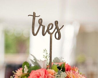 May include: A wooden table number sign with the word "tres" written in cursive, placed in a bouquet of pink and peach flowers.