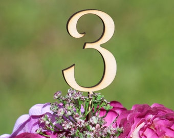 May include: A wooden number three table number topper, placed on a bouquet of pink and purple flowers.