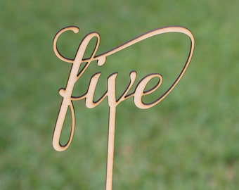 May include: A wooden table number with the word "five" written in a cursive font. The number is attached to a wooden stick.