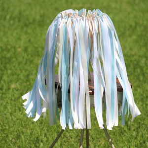 May include: A wedding ceremony ribbon wand with white and light blue ribbons attached to a wooden dowel. The wand is standing on a wooden base with three metal legs.