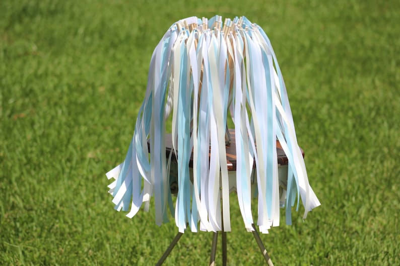 May include: A wedding ceremony ribbon wand with white and light blue ribbons attached to a wooden dowel. The wand is standing on a wooden base with three metal legs.