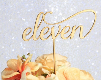 May include: A wooden cake topper with the word "eleven" in a cursive font. The topper is light brown and is placed on top of a bouquet of peach and orange flowers. The background is a sparkly white.
