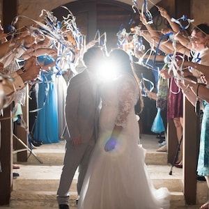 May include: A bride and groom kiss under a shower of blue and white ribbons as guests wave them overhead. The couple is standing on a stone staircase, and the bride is wearing a white wedding dress.