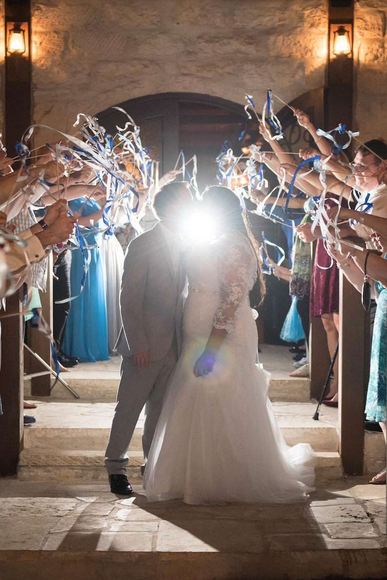 May include: A bride and groom kiss under a shower of blue and white ribbons as guests wave them overhead. The couple is standing on a stone staircase, and the bride is wearing a white wedding dress.
