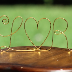 Könnte beinhalten: Goldene Drahtskulptur der Buchstaben "L" und "R" mit einem Herzen dazwischen. Die Skulptur steht auf einer Holzoberfläche.
