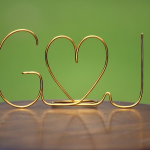 May include: A gold wire sculpture spelling out the letters "G & J" with a heart symbol in the middle. The sculpture sits on a dark wooden surface against a blurred green background. The letters are connected and stand upright.