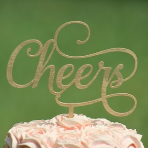 May include: A pink frosted cake with a gold "Cheers" topper.