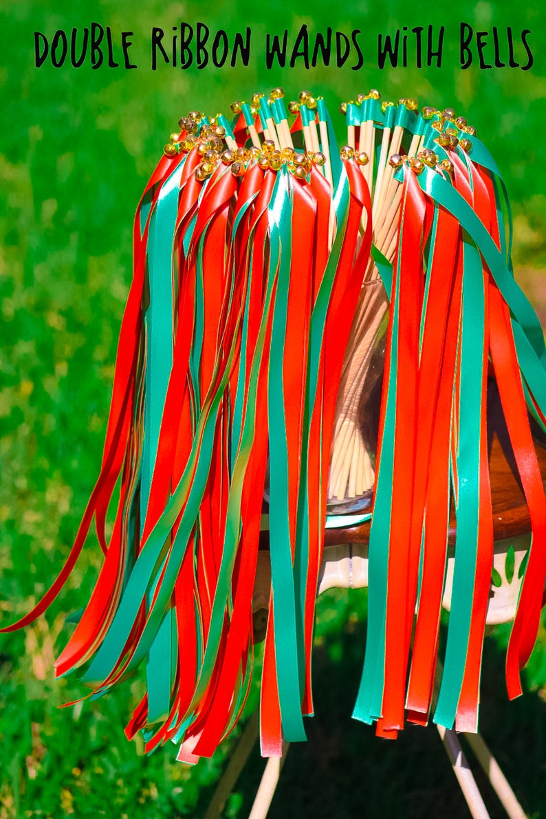 May include: A bunch of red and teal ribbon wands with gold bells at the top. The wands are tied together and are sitting on a white chair.