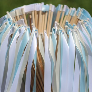 May include: A bundle of white and light blue ribbon wands, tied together at the top with white ribbon. The wands are made of wooden dowels with white ribbon tied around the top. The ribbons are flowing down from the top of the bundle.