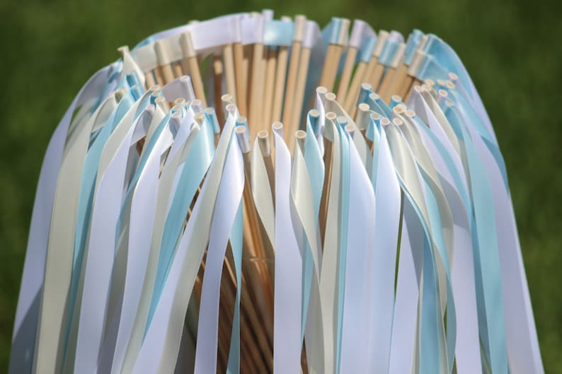 May include: A bundle of white and light blue ribbon wands, tied together at the top with white ribbon. The wands are made of wooden dowels with white ribbon tied around the top. The ribbons are flowing down from the top of the bundle.