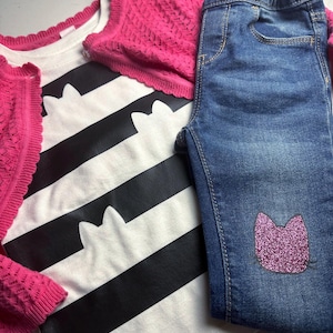 Może przedstawiać: Zestaw ubrań dla dzieci, w tym biało-czarna koszulka w paski z sylwetkami kotów, różowy kardigan i niebieskie dżinsy skinny z różową, brokatową naszywką w kształcie kota. Strój jest ułożony na jasnoszarej powierzchni.