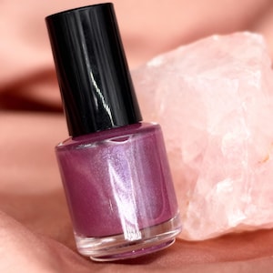 Puede incluir: Un frasco de esmalte de uñas brillante de color ciruela con una tapa negra. El frasco de esmalte de uñas se coloca sobre un fondo rosa suave y un cristal rosa claro. El esmalte de uñas tiene un frasco de vidrio transparente.