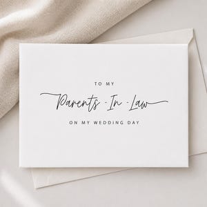 May include: A white card with the text "TO MY Parents-In-Law ON MY WEDDING DAY" in elegant black script. The card is placed on a white envelope, with a neutral-colored fabric in the background.