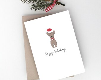 May include: A white card with a brown envelope. The card has a cartoon illustration of a brown dog wearing a red Santa hat and a red collar with a heart. The text "happy holidays!" is written in black cursive below the dog.