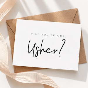 May include: A white rectangular greeting card with black text that reads Will You Be Our Usher? The card is placed on top of a brown kraft paper envelope, accompanied by a light beige ribbon that curves across the bottom of the composition.
