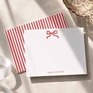 May include: A stationery set featuring a white notepad with a red bow illustration and the name "sophia elizabeth" printed in red. A red and white striped notepad is behind it. A white ribbon is on the left.