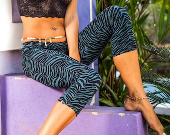 May include: Two women wearing patterned leggings and tops pose in front of a wooden structure. The woman on the left is wearing a black top with a blue and black patterned leggings. The woman on the right is wearing a black lace top with a blue and black zebra patterned leggings. The background features a wooden structure and lush green foliage.