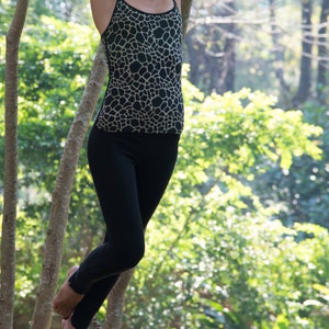 May include: A woman wearing a black and tan patterned tank top and black leggings, hanging from tree branches in a forest setting. The tank top has a halter neckline and a giraffe-like pattern. The leggings are form-fitting.