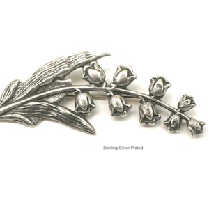 May include: A silver-plated brooch in the shape of a lily of the valley. The brooch features a detailed leaf and a stem with several bell-shaped flowers. The text "Sterling Silver Plated" is visible.