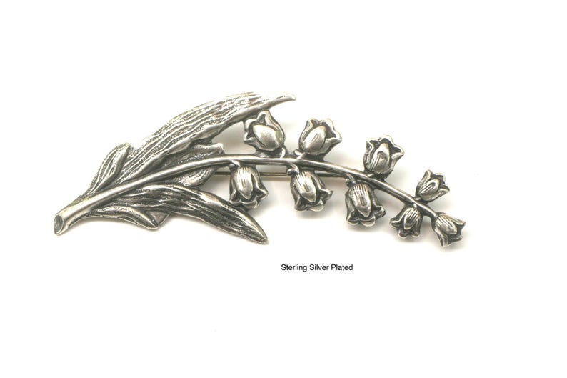 May include: A silver-plated brooch in the shape of a lily of the valley. The brooch features a detailed leaf and a stem with several bell-shaped flowers. The text "Sterling Silver Plated" is visible.