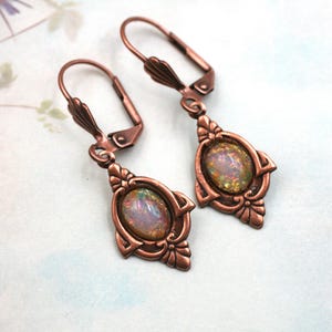 May include: A pair of copper-colored drop earrings. Each earring features an oval, iridescent stone set within a decorative frame. The earrings have a lever-back closure and an ornate, vintage design.